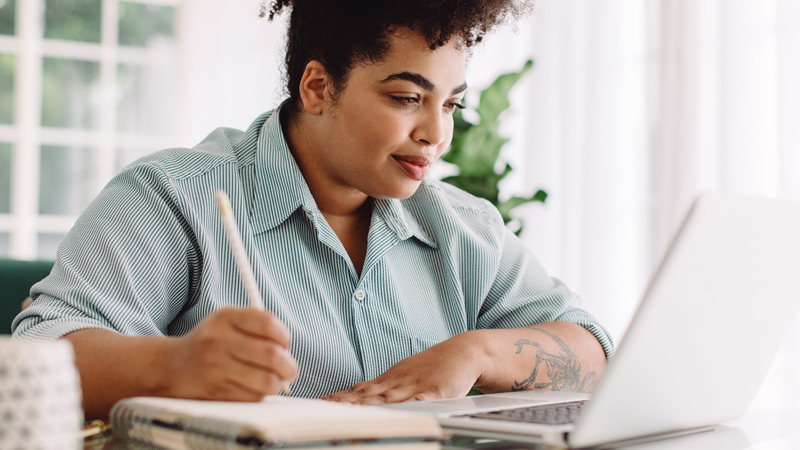 a woman taking notes on pen and paper while looking at her laptop