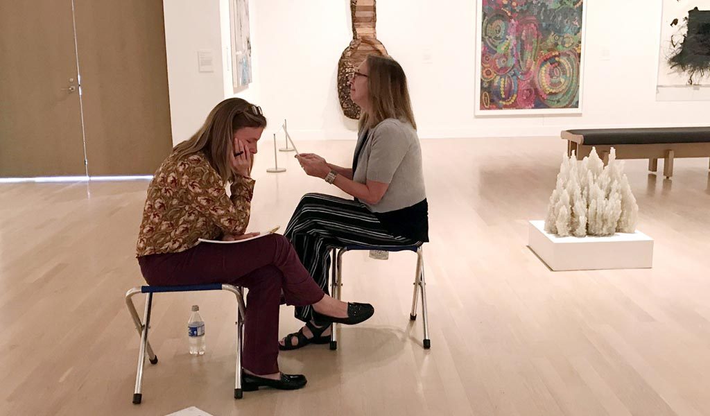 two women sit in a gallery taking notes 