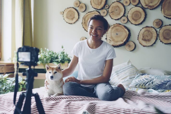 a woman filming a video of her self talking to the camera while sitting on the bed with her dog