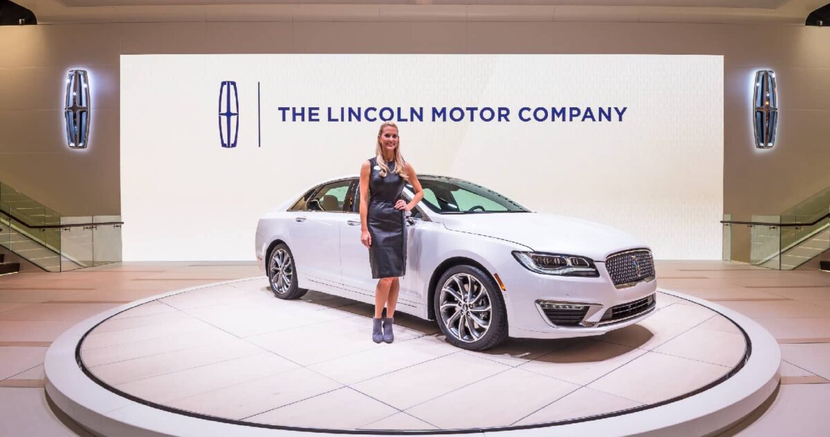 A white Lincoln sedan in a showroom with a woman standing in front of the vehicle. She is wearing a black cocktail dress and her left hand is on her hip. Behind the car is a wall with "The Lincoln Motor Company" illuminated.
