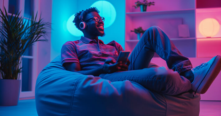 Black man with glasses sitting on a bean bag chair, wearing headphones and smiling.