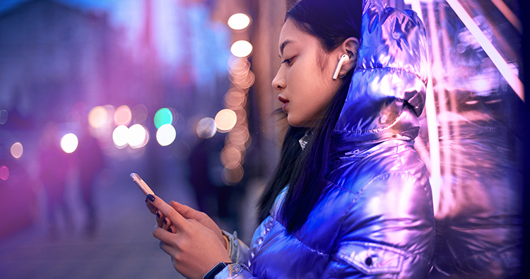 Side view of trendy teenager girl in glowing neon light standing on street, using online app on modern smartphone to listen to audio content