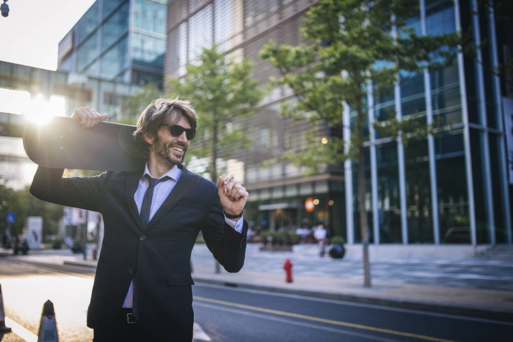 Man wearing a suit and tie with sunglasses, holding a skateboard over his shoulder. He is standing on the street in front of a brick and glass building. The sun is shining behind him.