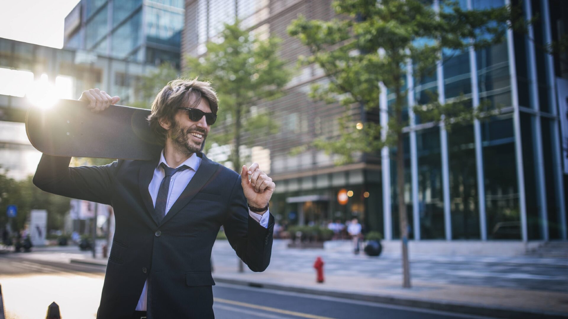 Man wearing a suit and tie with sunglasses, holding a skateboard over his shoulder. He is standing on the street in front of a brick and glass building. The sun is shining behind him.