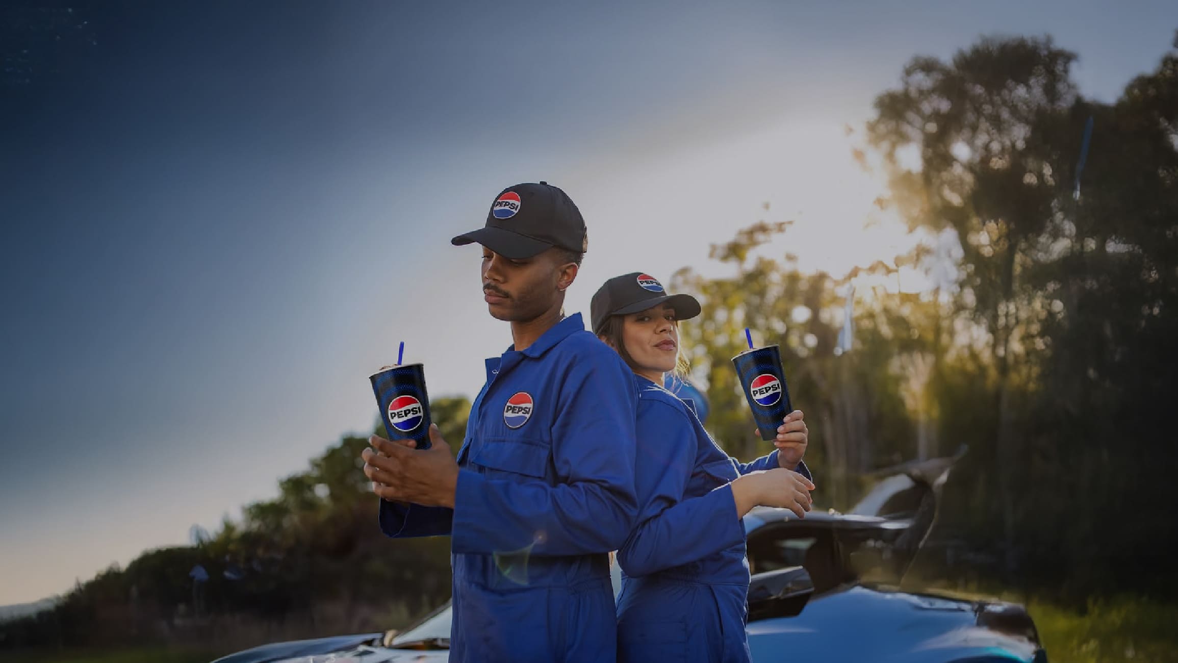 A man and a woman standing back to back wearing Pepsi branded jumpsuits, ball caps, and holding Pepsi drink cups.